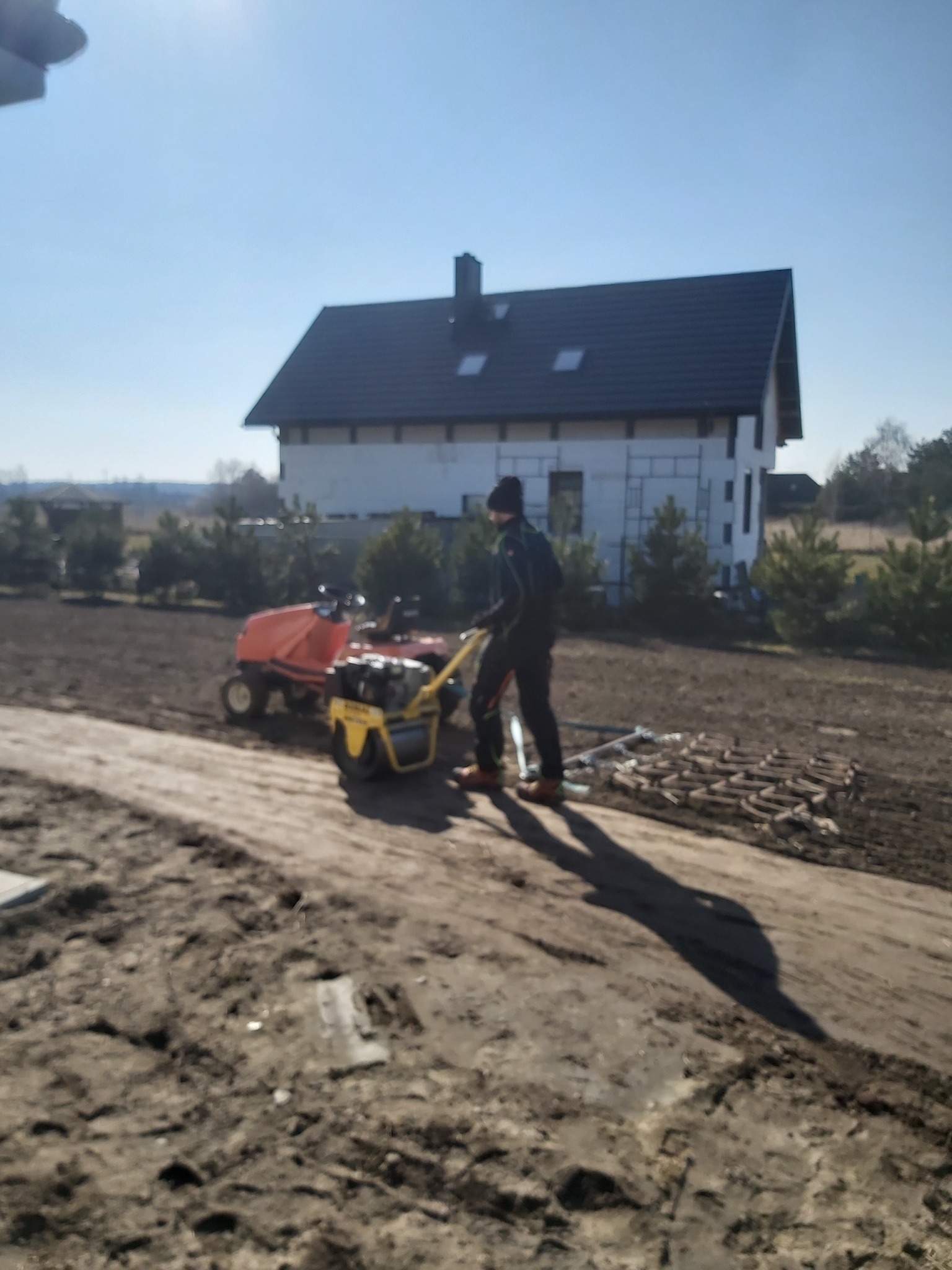 Pracownik w ciemnej odzieży wyrównuje glebę za pomocą glebogryzarki na działce przed domem w trakcie budowy, w tle widoczne drzewa i niebo.
