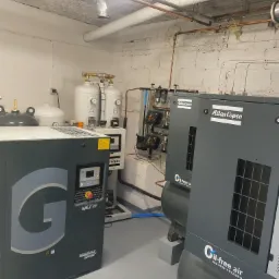 Pomieszczenie z dwoma szarymi kompresorami Atlas Copco 'Oil-free air', zbiornikami ciśnieniowymi z manometrami oraz instalacją rur pod sufitem z betonową fakturą.