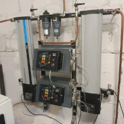 Kompletny system uzdatniania sprężonego powietrza Atlas Copco CD3+ z filtrami POP10+ i miedzianą instalacją rurową, zamontowany na białej, ceglanej ścianie w piwnicy.