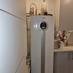 Biała pompa ciepła IVT z widocznymi rurami i manometrem, umieszczona w kuchni z białymi i szarymi szafkami, obok drzwi z widocznymi kablami.