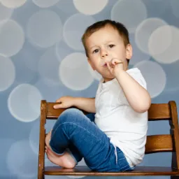 Chłopiec w białej koszulce i dżinsach siedzi na drewnianym krześle na tle rozmytych światełek, trzymając w ustach mały patyczek.