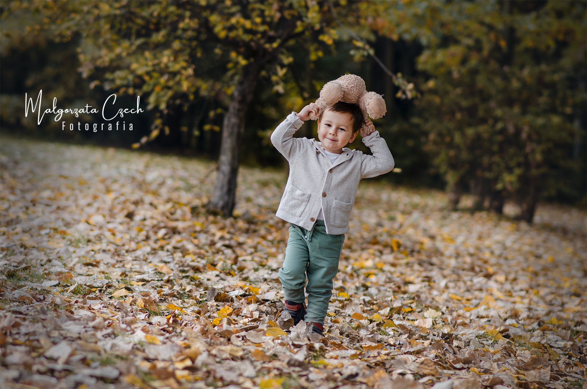 Uśmiechnięty chłopiec w jesiennej scenerii trzyma pluszowego misia nad głową, otoczony opadłymi liśćmi.
