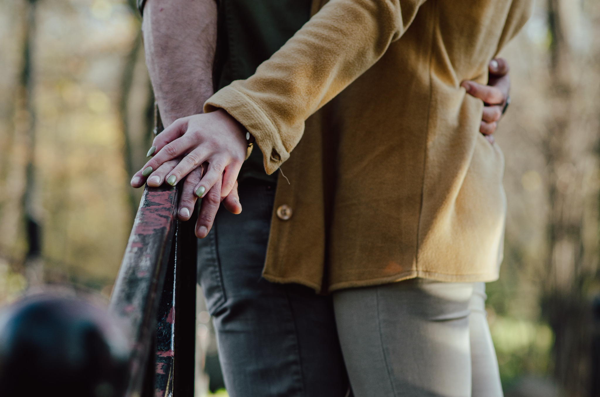 Para obejmująca się na tle drzew, dłonie kobiety z zielonym manicure spoczywają na ręce mężczyzny na metalowej barierce.