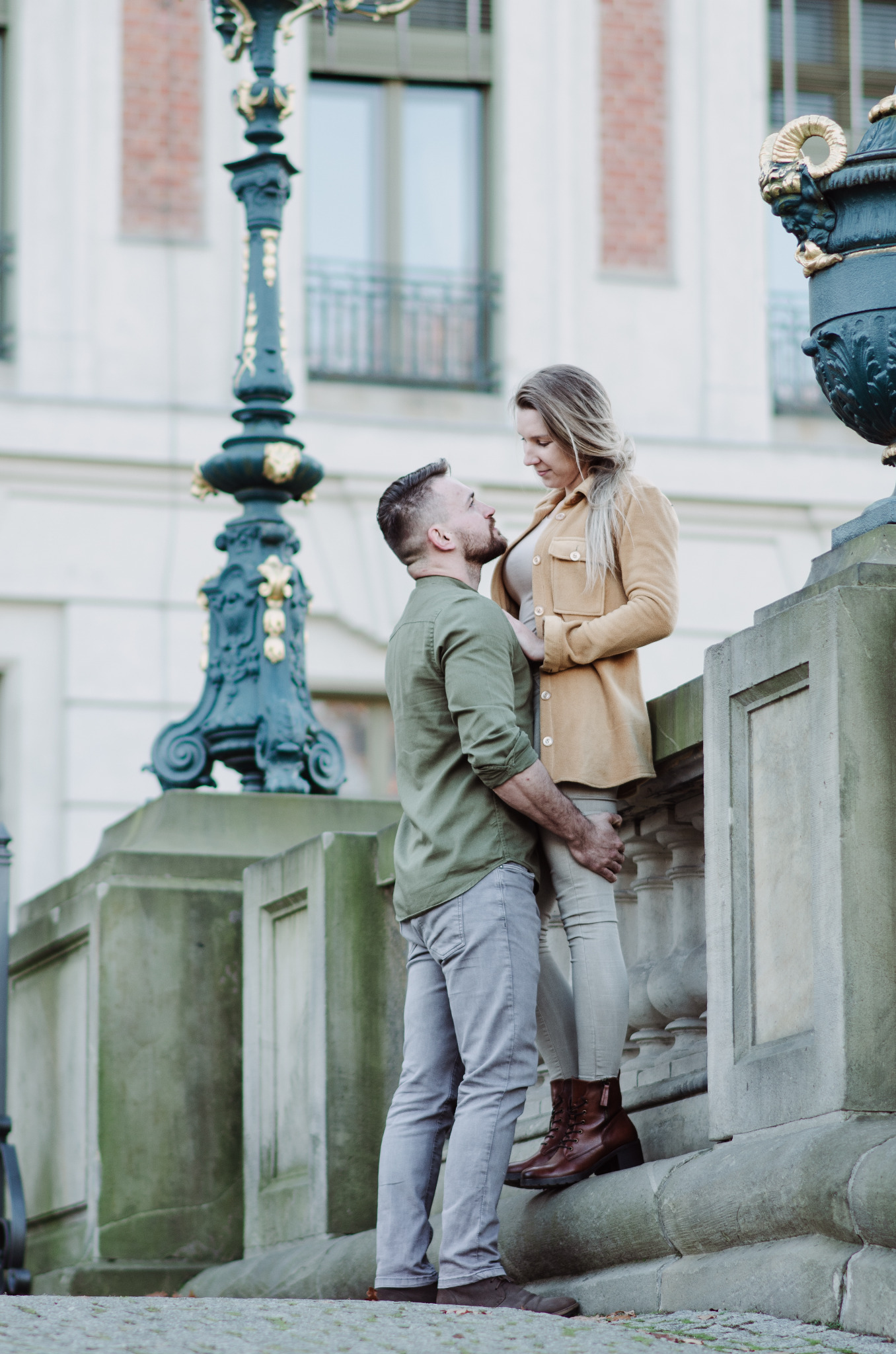 Para zakochanych na tle klasycznej architektury miejskiej; mężczyzna w zielonej koszuli i szarych spodniach obejmuje kobietę w beżowej kurtce i brązowych botkach.