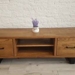 Drewniana szafka RTV z dwoma szufladami i półką, wykończona w ciepłym odcieniu brązu, z czarnymi uchwytami i nóżkami, na blacie doniczka z lawendą, tło stanowi ściana z białej cegły.