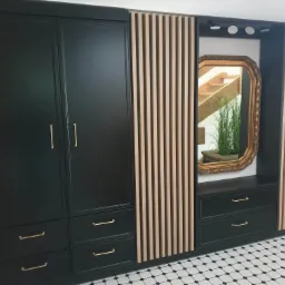 Czarna zabudowa meblowa z szafami, szufladami, lustrem w ozdobnej ramie i dekoracyjnymi panelami z drewnianych listew, na podłodze wzór szachownicy.