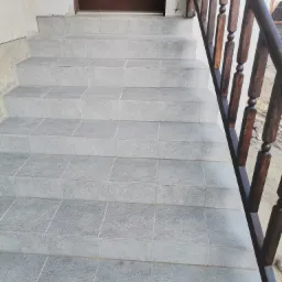 Schody wejściowe wyłożone szarą, kwadratową płytką, z ciemnobrązową drewnianą balustradą po prawej stronie, prowadzące do brązowych drzwi wejściowych.