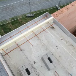 Przygotowanie zbrojenia stropu betonowego z użyciem prętów i styropianu, widok z góry na plac budowy z ogrodzeniem tymczasowym w tle.