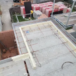 Widok z góry na strop budynku w trakcie budowy: betonowe płyty, zbrojenie stalowe, obrzeża z izolacji termicznej, w tle cegły Porotherm, rusztowanie i żuraw budowlany.