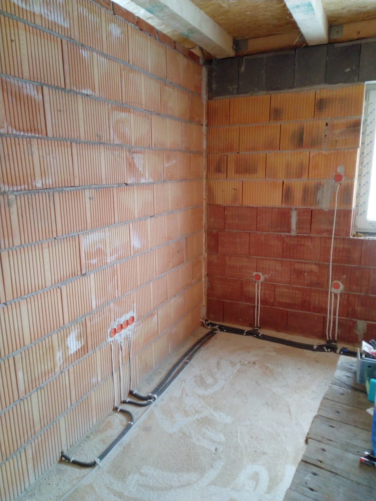 Wnętrze w trakcie prac instalacyjnych: ściany z cegły ceramicznej z widocznymi przewodami elektrycznymi w peszlach, puszki instalacyjne w ścianie, surowy beton na podłodze.