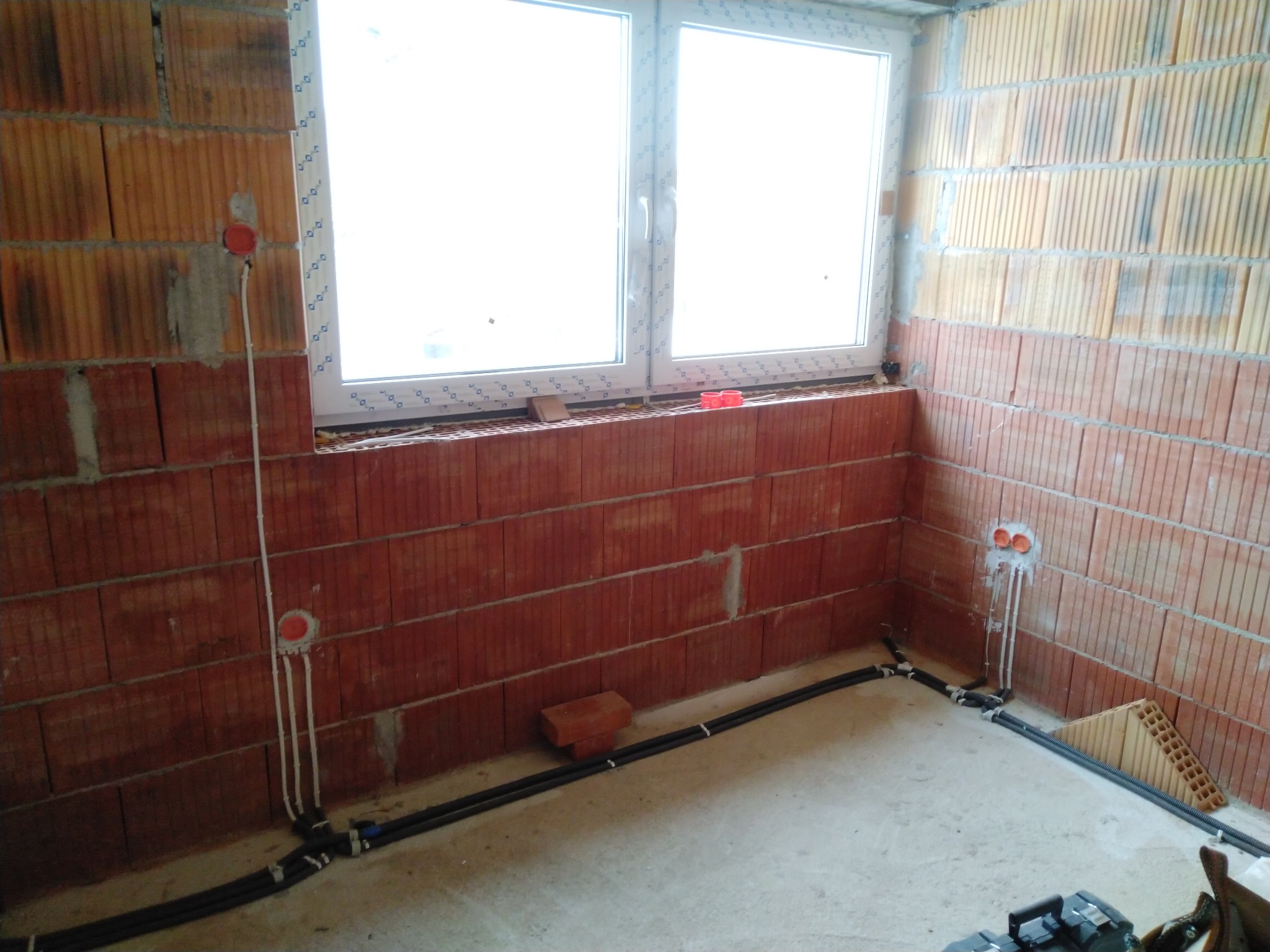 Wewnętrzne okablowanie elektryczne w trakcie instalacji w surowym pomieszczeniu z ceglanymi ścianami i oknem; widoczne puszki instalacyjne i peszle.
