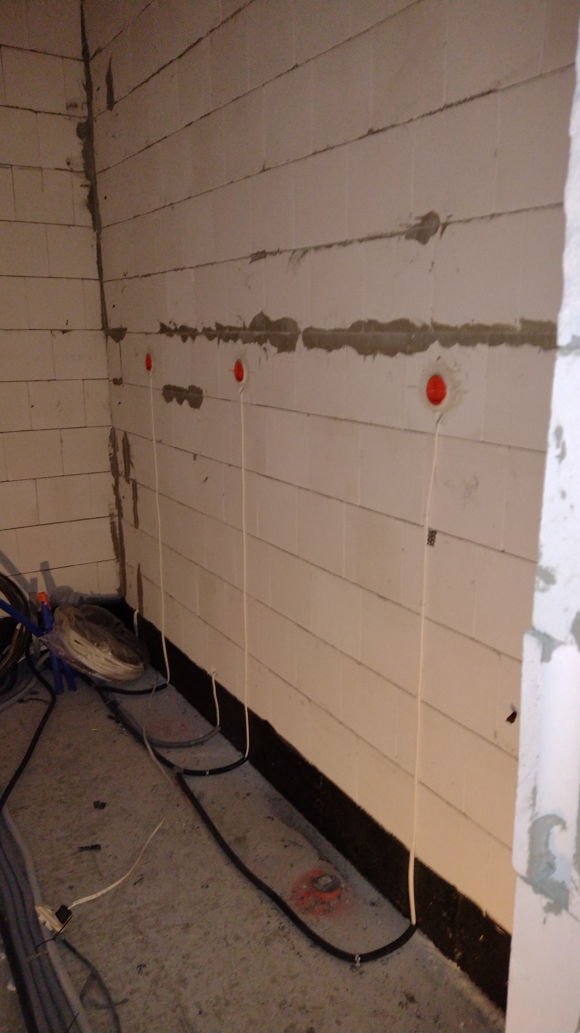 Wewnętrzne ściany z pustaków z widocznymi przewodami elektrycznymi wychodzącymi z czerwonych puszek instalacyjnych, ułożone na podłodze kable w czarnej izolacji, przygotowanie do instalacji...