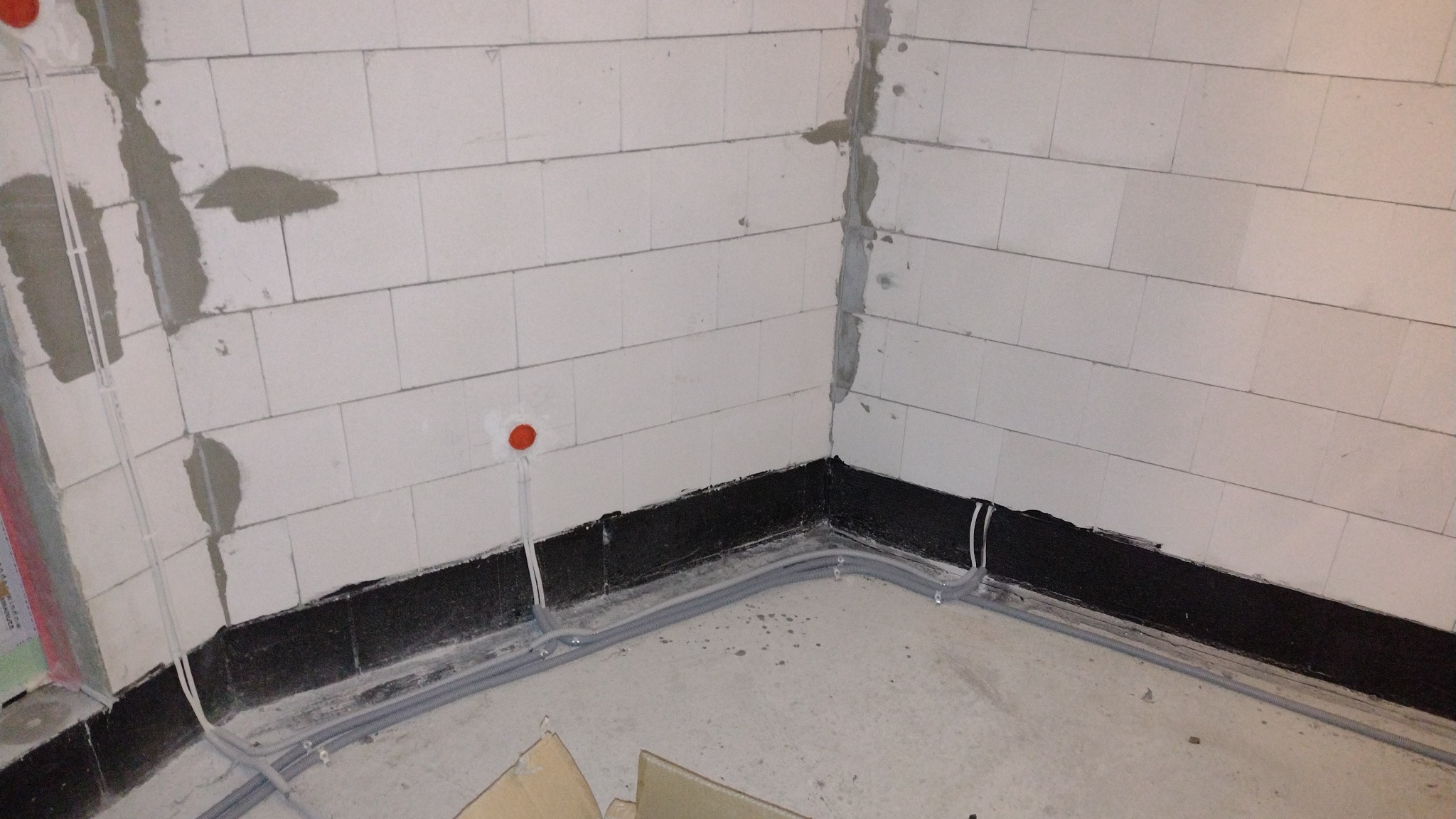 Wrocław: Instalacja elektryczna w surowym stanie, widoczne kable w peszlach prowadzone wzdłuż ściany z bloczków, puszka elektryczna w kolorze pomarańczowym.