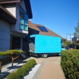 PioMat transport i przeprowadzki - Jasnoniebieski samochód dostawczy z otwartą przestrzenią ładunkową zaparkowany przed nowoczesnym domem z panelami słonecznymi na dachu, widoczne przedmioty przygotowane do załadunku.