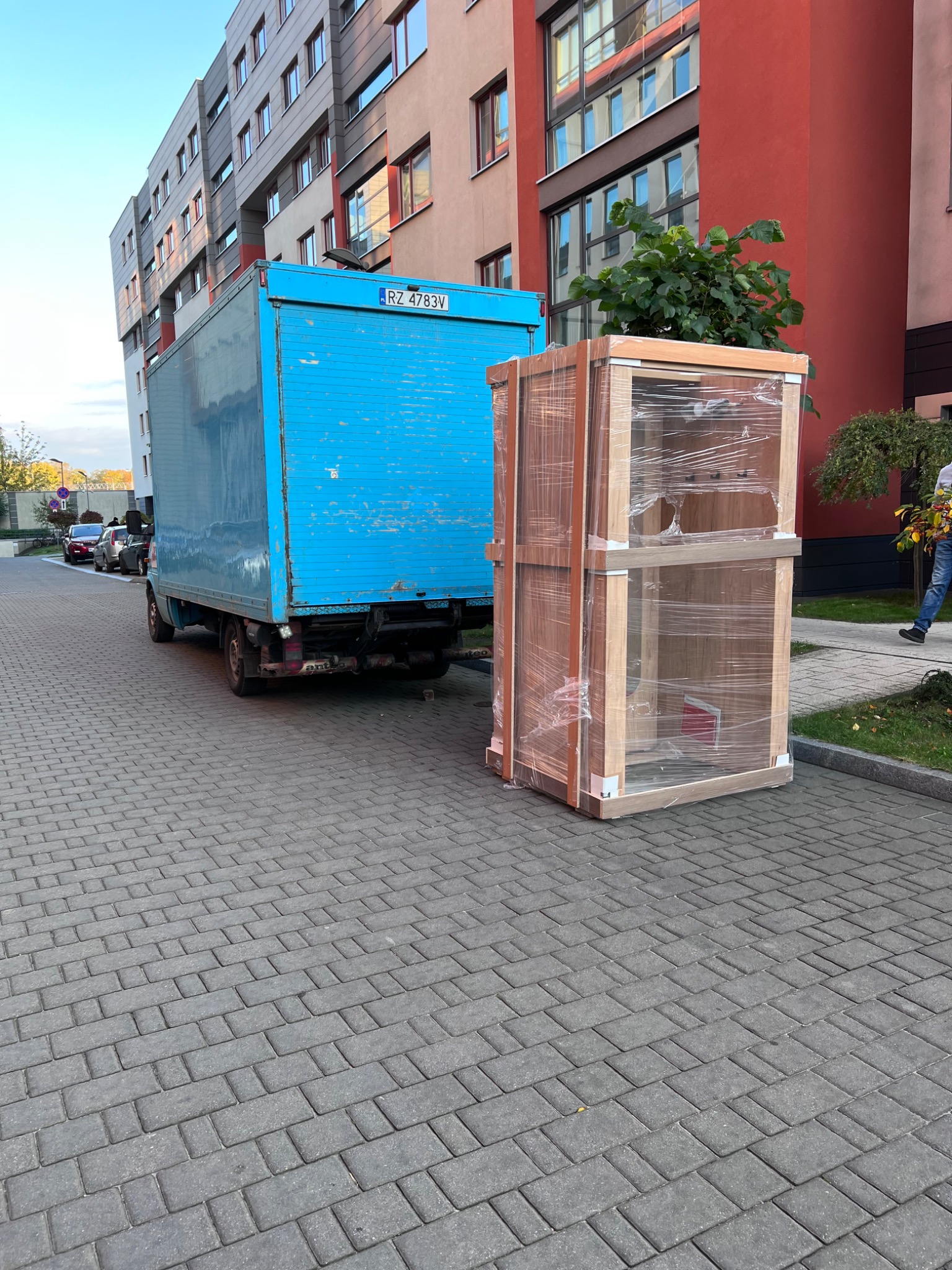 Niebieski samochód dostawczy z otwartą przestrzenią ładunkową stoi na kostce brukowej obok zapakowanego w folię mebla z jasnego drewna na tle nowoczesnego bloku mieszkalnego.