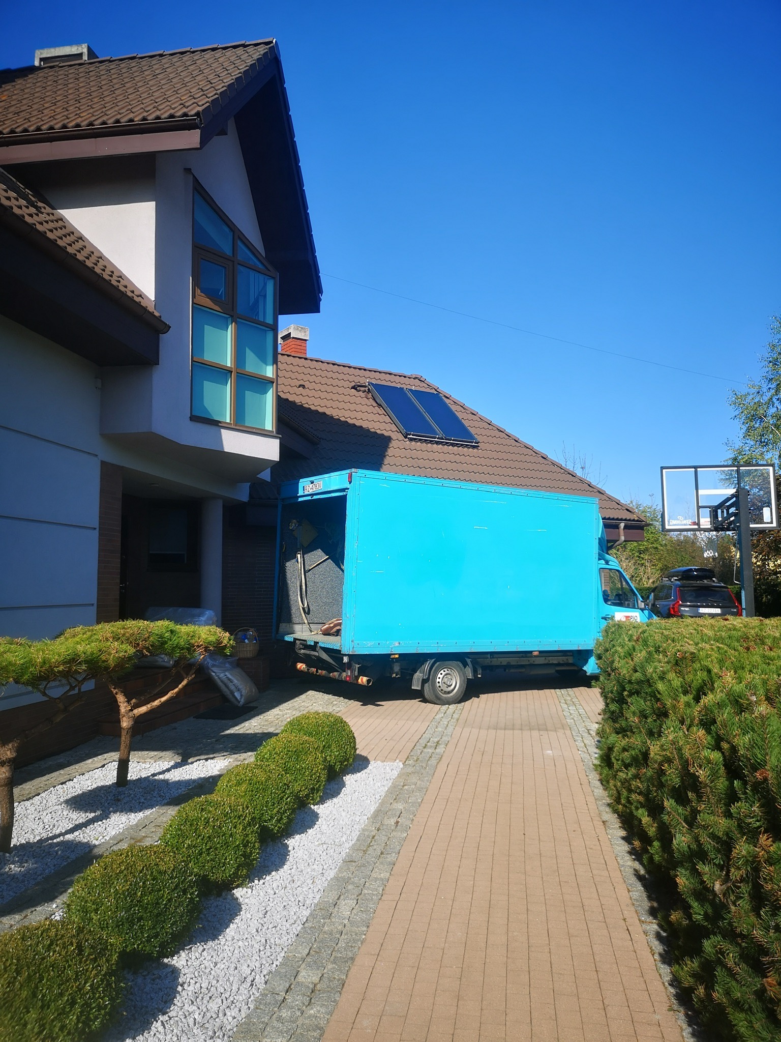 Jasnoniebieski samochód dostawczy z otwartą przestrzenią ładunkową zaparkowany przed nowoczesnym domem z panelami słonecznymi na dachu, widoczne przedmioty przygotowane do załadunku.