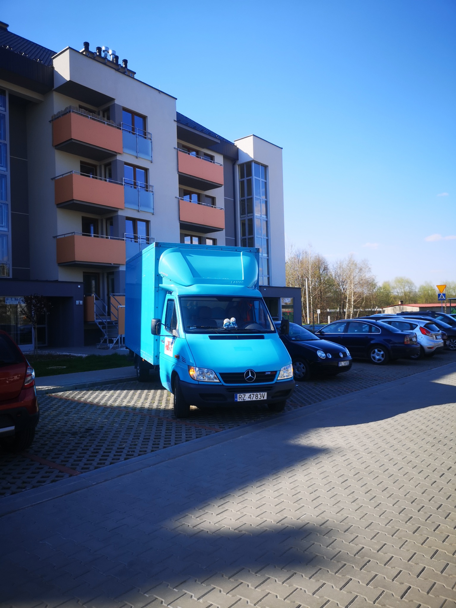 Niebieski samochód dostawczy zaparkowany przed nowoczesnym budynkiem mieszkalnym z pomarańczowymi balkonami, w tle widoczne inne zaparkowane samochody.