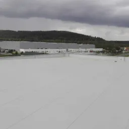 Rozległy, płaski dach pokryty jasną membraną, widok z góry na budynek przemysłowy w tle i zachmurzone niebo.