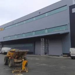 Nowoczesny budynek przemysłowy z szarej płyty warstwowej, z widocznym logo firmy 'sikla', rusztowaniem i mini-ładowarką na paletach przed budynkiem.