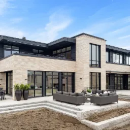 Nowoczesny dwupiętrowy dom z jasnej cegły klinkierowej, czarne ramy okienne, taras z szarymi meblami ogrodowymi i donicami z roślinami, minimalistyczny design.