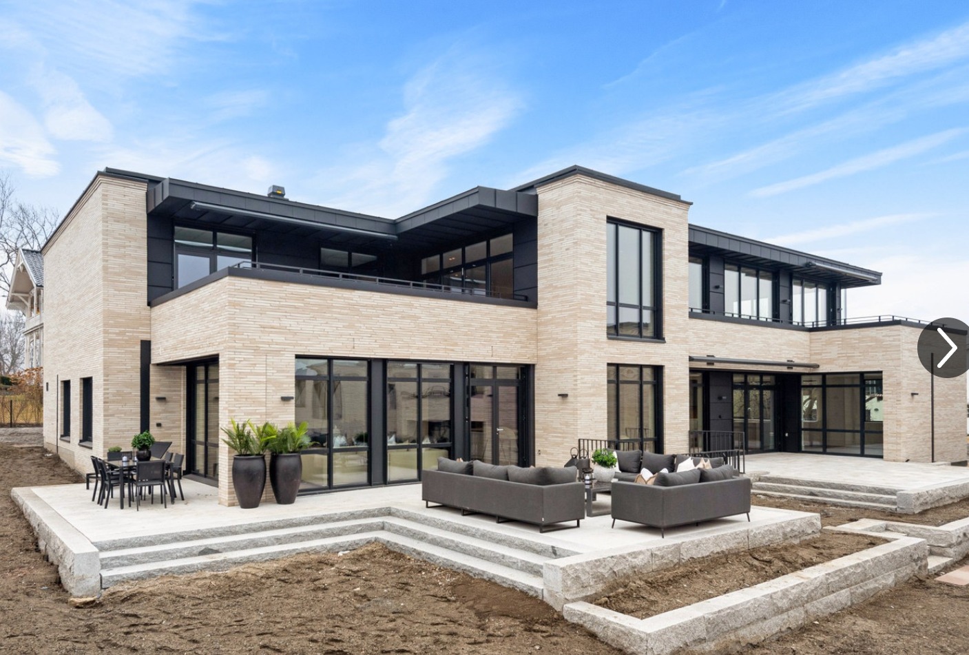 Nowoczesny dwupiętrowy dom z jasnej cegły klinkierowej, czarne ramy okienne, taras z szarymi meblami ogrodowymi i donicami z roślinami, minimalistyczny design.