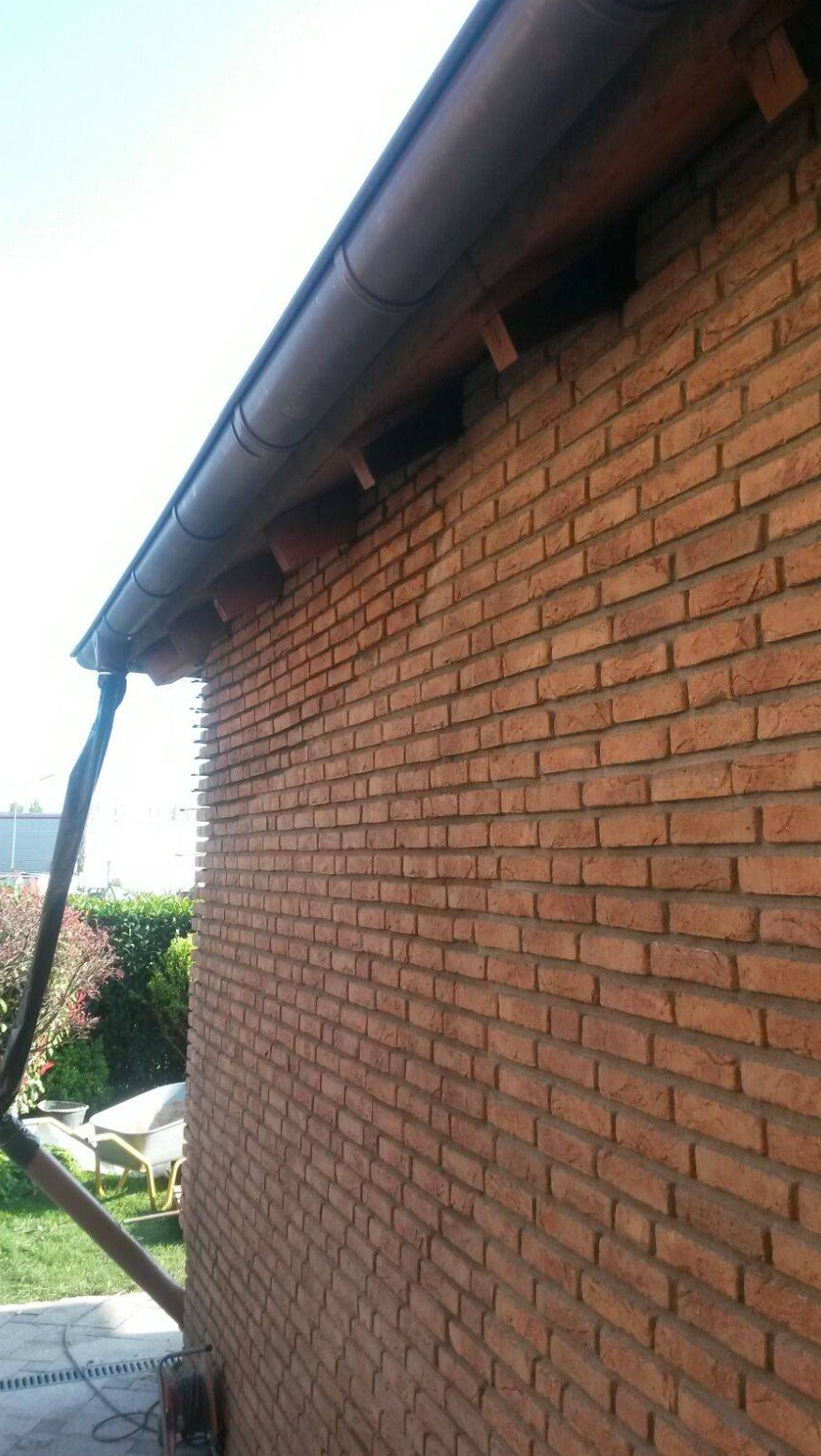 Ściana elewacyjna z cegły klinkierowej pod brązowym orynnowaniem, widoczny wózek budowlany i fragment trawnika.