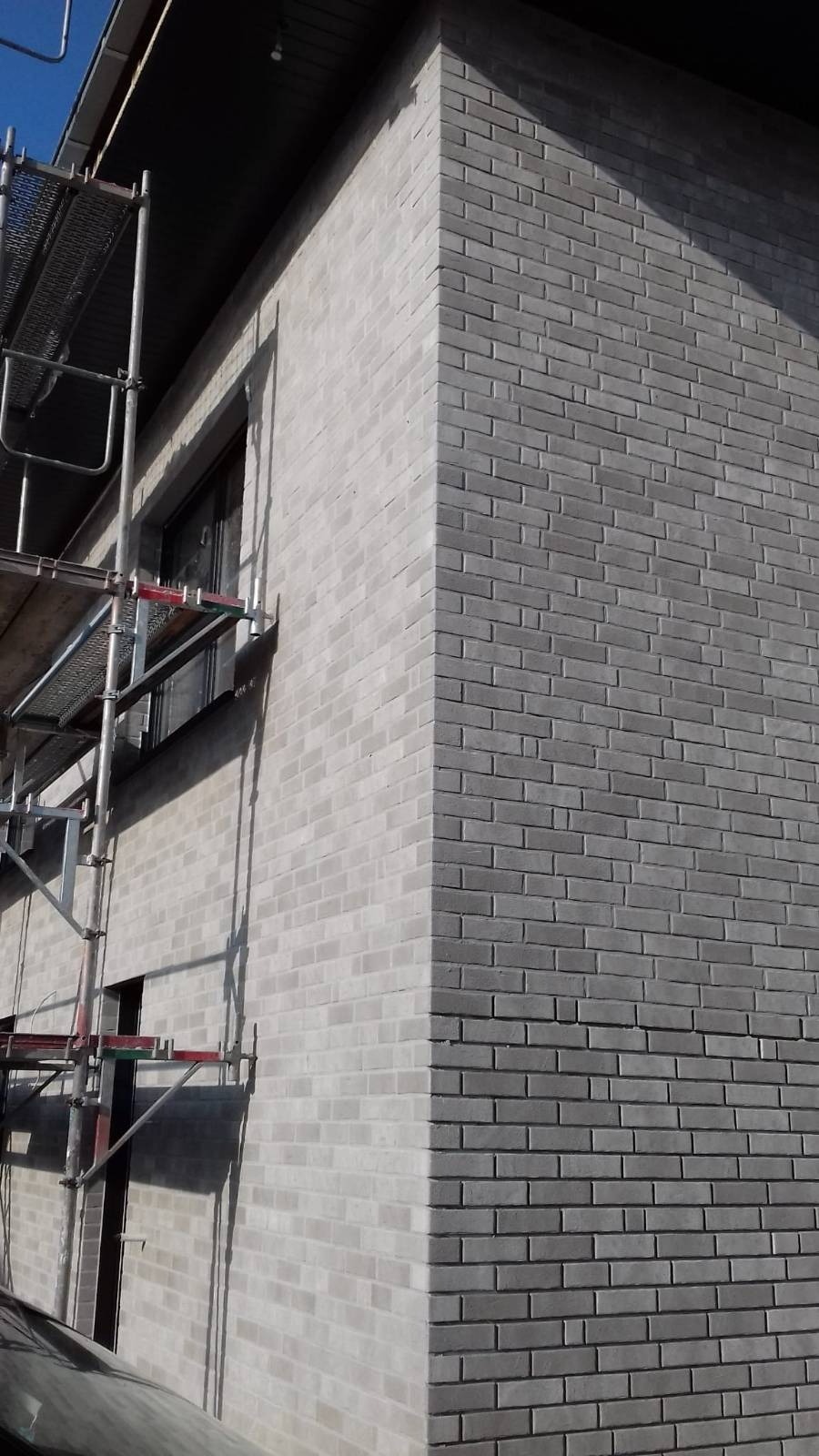 Szara ceglana elewacja budynku z rusztowaniem, widoczny narożnik i fragment okna.