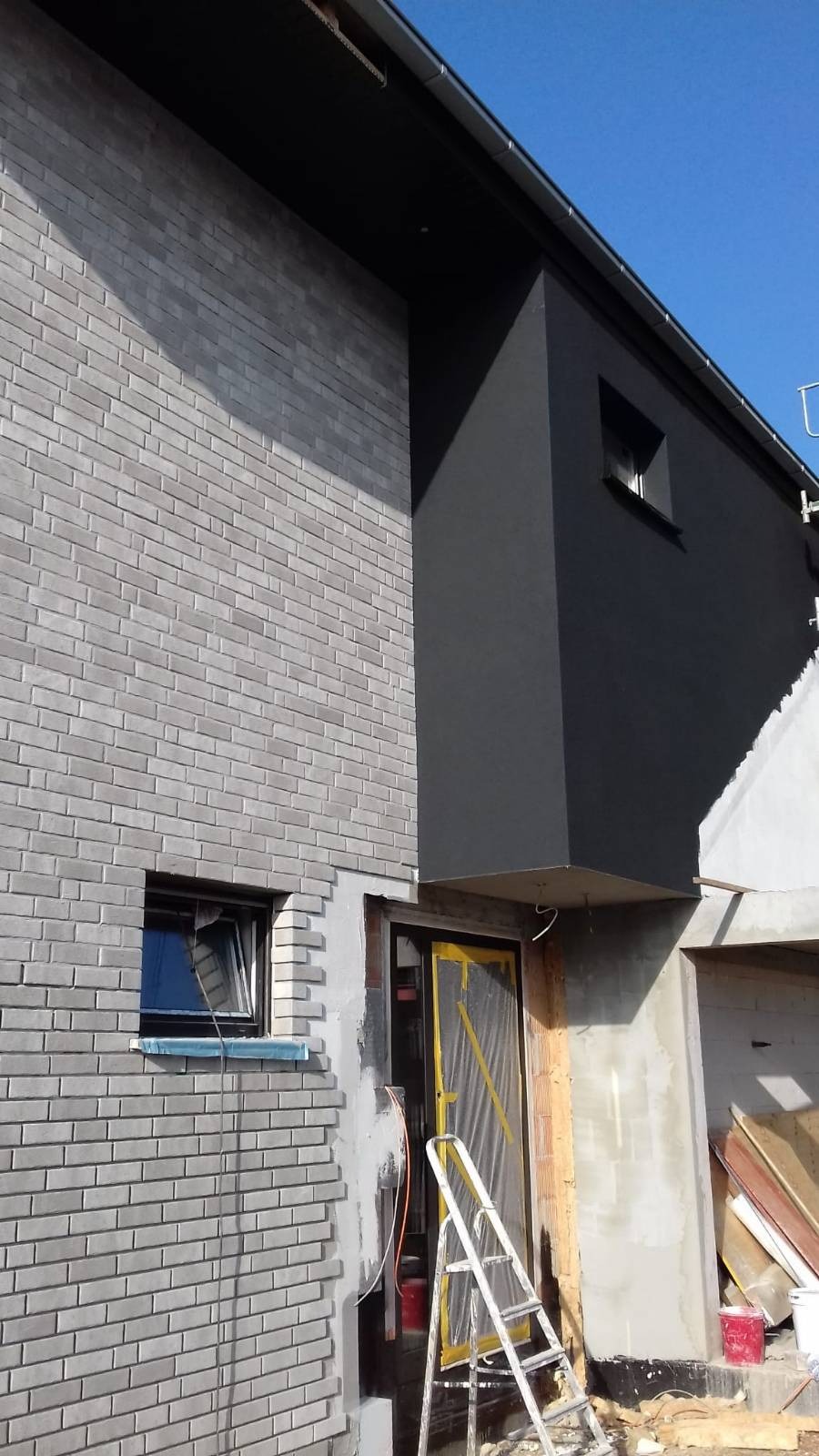 Dwukolorowa elewacja domu w trakcie prac wykończeniowych: szara cegła klinkierowa i czarny tynk, drabina oparta o zabezpieczone folią drzwi wejściowe.