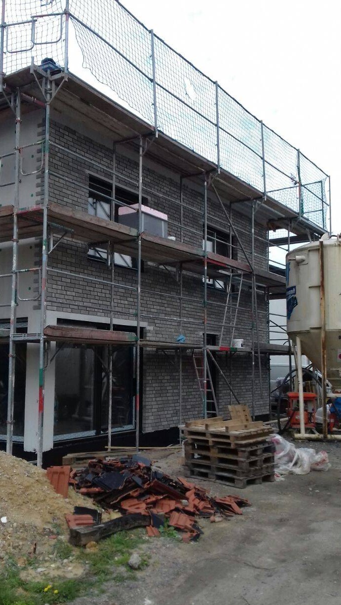 Budynek w trakcie prac elewacyjnych, widoczne rusztowania z siatką ochronną, częściowo ocieplona ściana z szarej cegły, palety i gruz dachówkowy na ziemi, oraz silos na zaprawę.