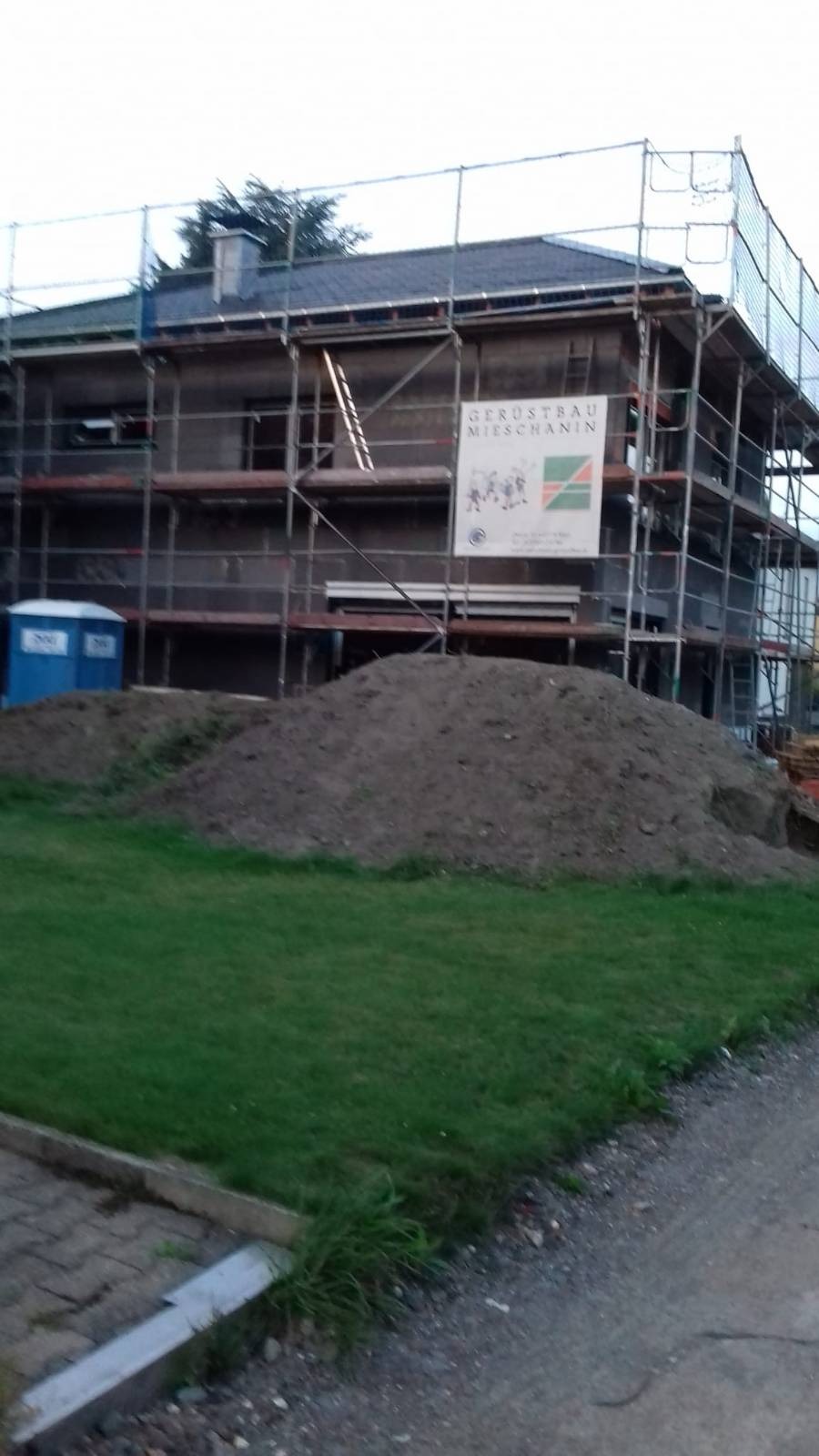 Dom w trakcie prac elewacyjnych, otoczony rusztowaniem, z widocznym banerem firmy budowlanej i stertą ziemi przed budynkiem.