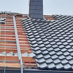 Fragment dachu w trakcie układania dachówki ceramicznej w kolorze grafitowym, widoczny komin obłożony łupkiem oraz elementy konstrukcji drewnianej i metalowe profile.