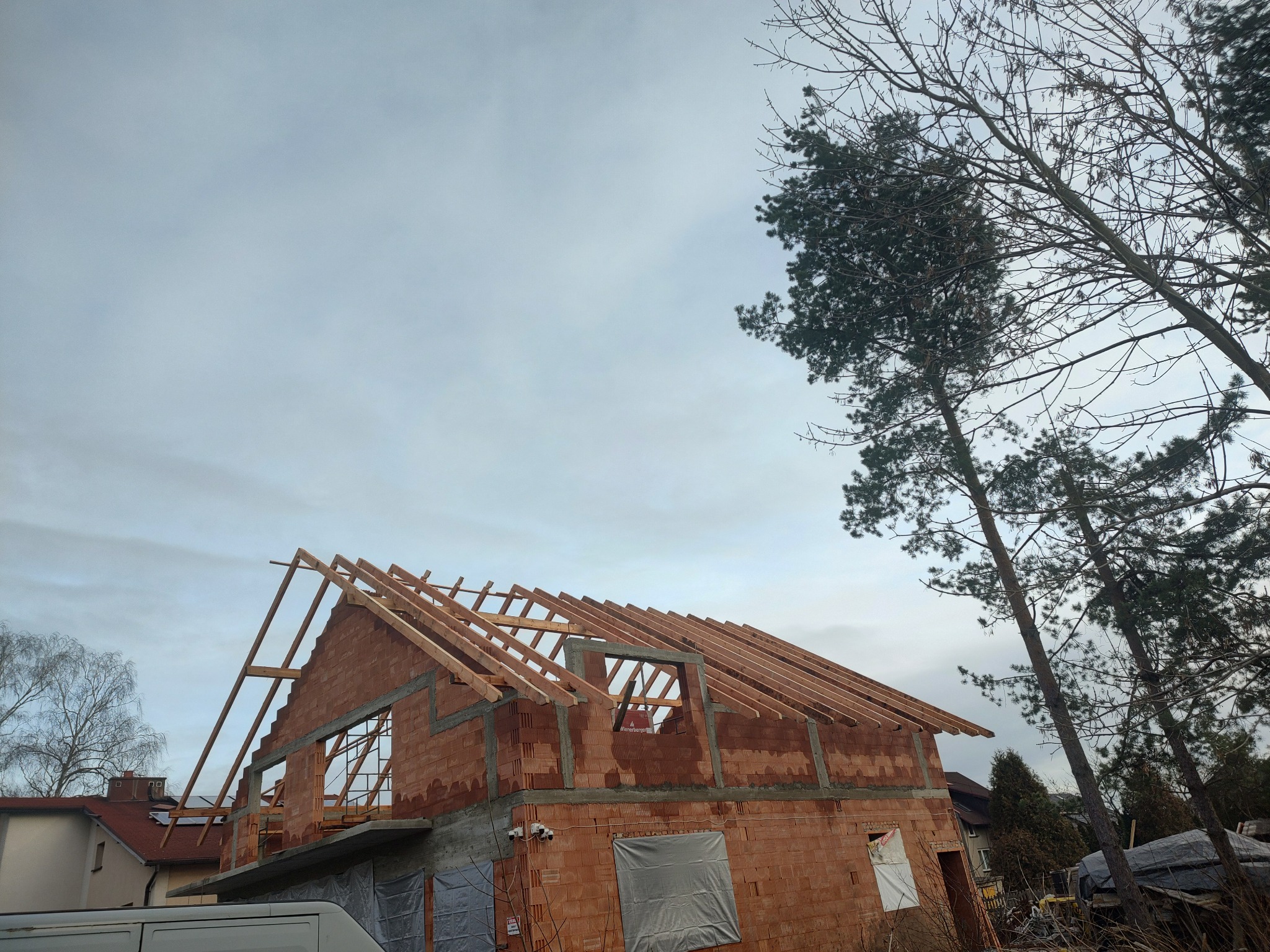Surowy stan budynku z czerwonej cegły w trakcie budowy, z widoczną drewnianą konstrukcją dachu i elementami deskowania, na tle pochmurnego nieba i drzew