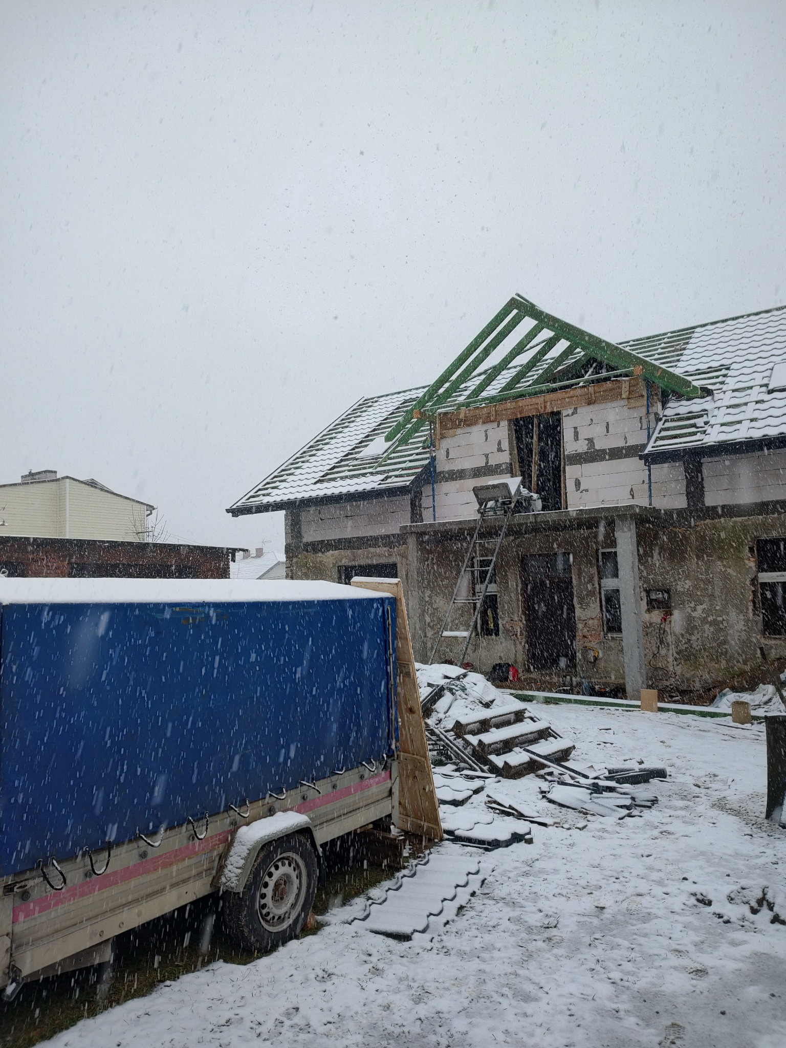 Niedokończony dom w trakcie budowy dachu podczas śnieżnej pogody, widoczna więźba dachowa, częściowo pokryty dachówką, przyczepa z plandeką na pierwszym planie.