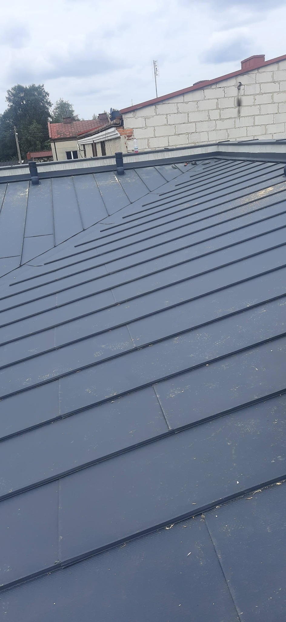 Nowoczesne, grafitowe pokrycie dachowe z blachy na ukończonym dachu, widoczne kominki wentylacyjne i fragment budynku w tle.