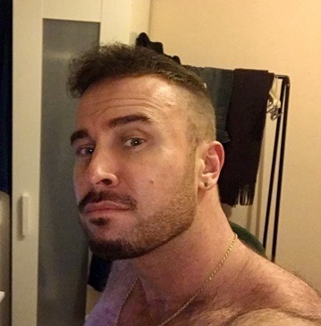 Mężczyzna o krótkich, kręconych włosach, z lekkim zarostem, w złotej biżuterii, pozujący do zdjęcia z lekkim uśmiechem.