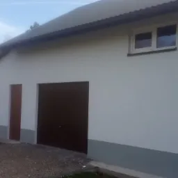 Świeżo pomalowana elewacja budynku gospodarczego w odcieniach bieli i szarości, z brązowymi drzwiami garażowymi i wejściowymi, na tle zieleni i domu mieszkalnego.