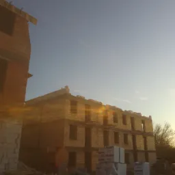 Wznoszony budynek z bloczków SOLBET, widoczne palety i materiały budowlane, w tle błękitne niebo, ciepłe światło zachodzącego słońca.