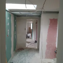 Wnętrze w trakcie prac wykończeniowych: widoczne profile sufitowe, częściowo położona płyta gipsowo-kartonowa na ścianach z widocznymi otworami na instalacje, przewody elektryczne zwisające...