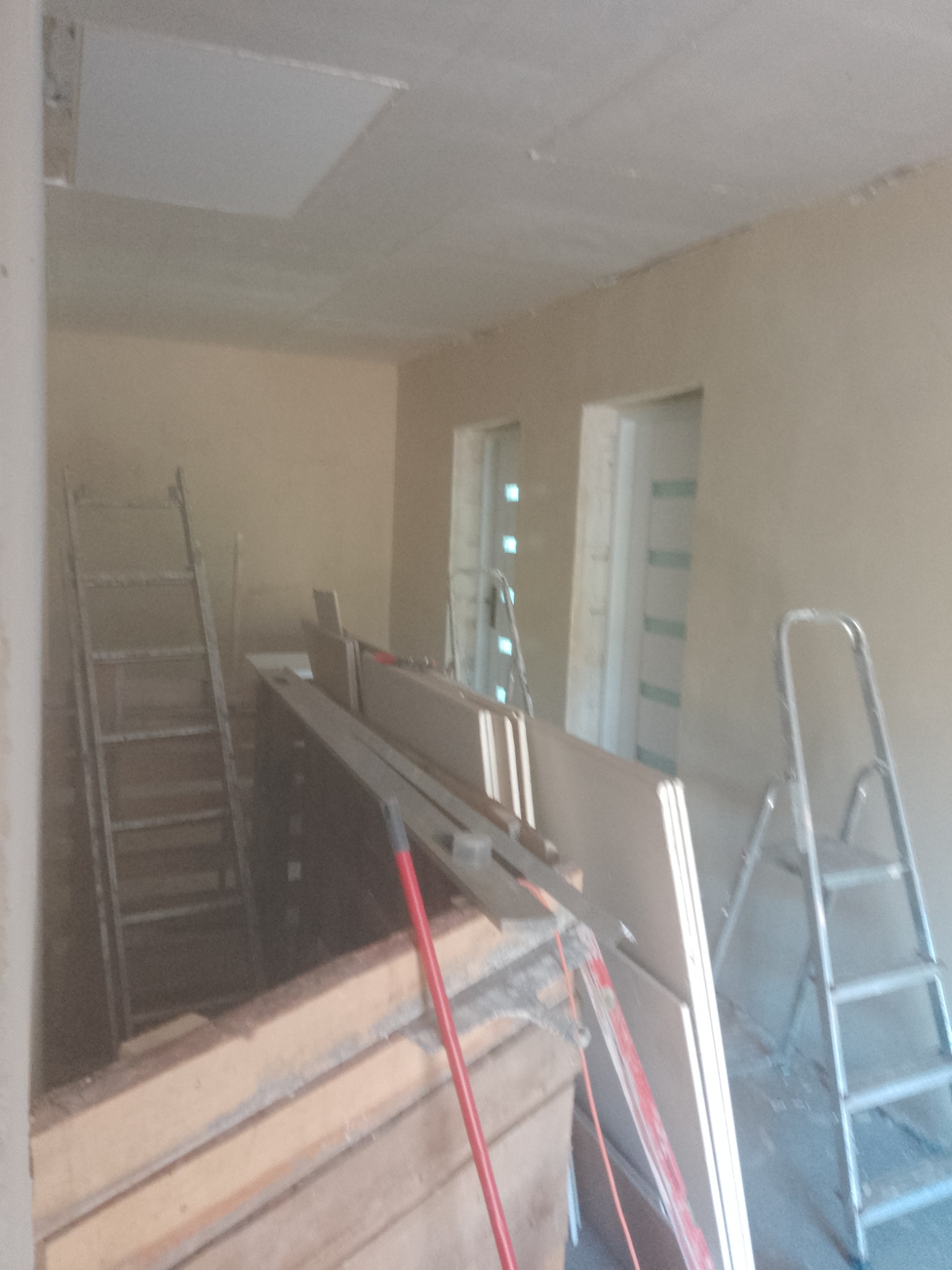 Wnętrze w trakcie prac wykończeniowych: surowe ściany, płyty karton-gips oparte o ścianę, dwie drabiny aluminiowe, drewniane belki i czerwony uchwyt narzędzia w nieostrym planie.