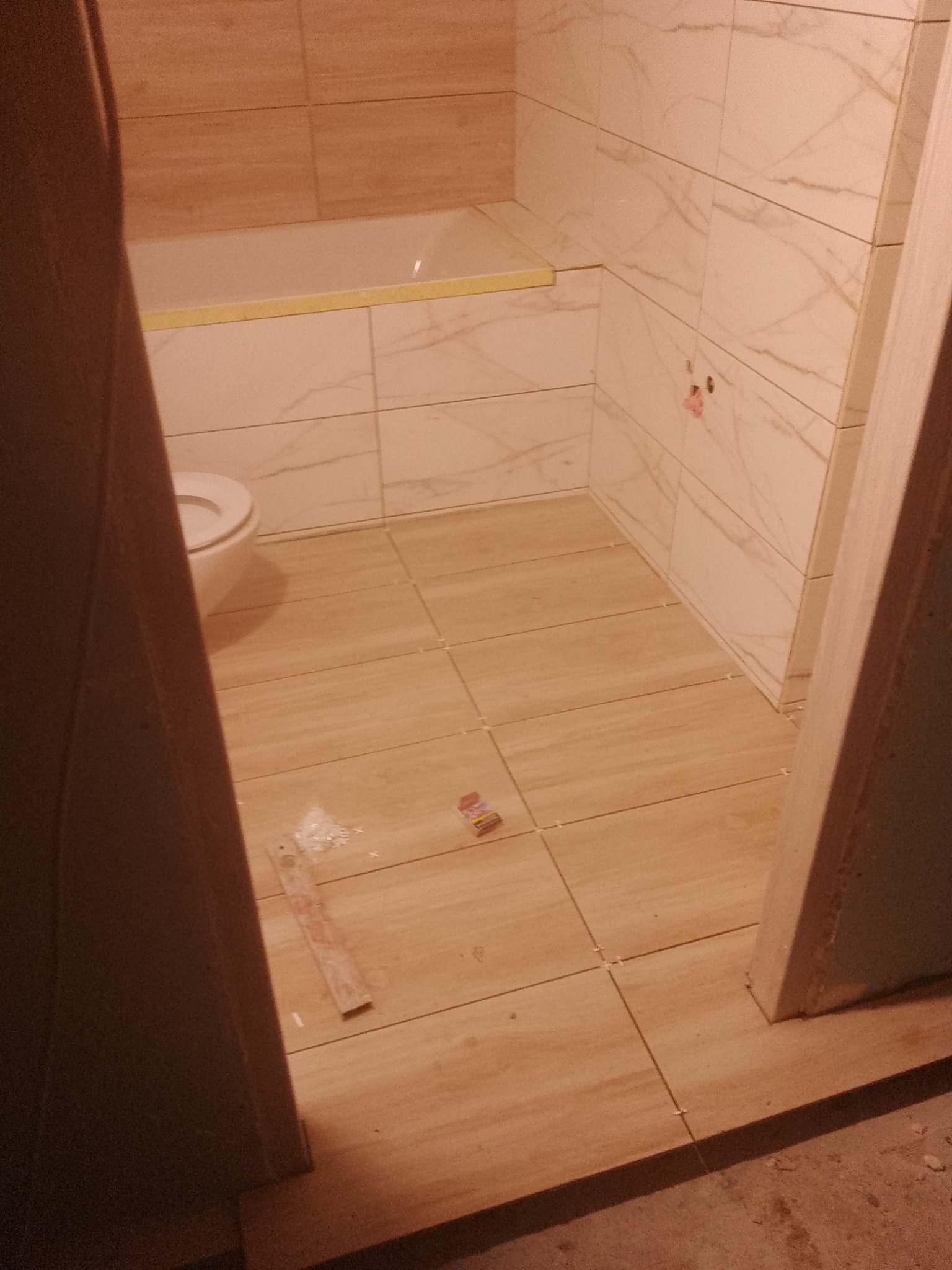 Wykończona łazienka z beżowymi płytkami imitującymi drewno na podłodze i marmur na ścianach, widoczna wanna obłożona płytkami i fragment toalety, z elementami narzędzi i pozostałościami po pracach...