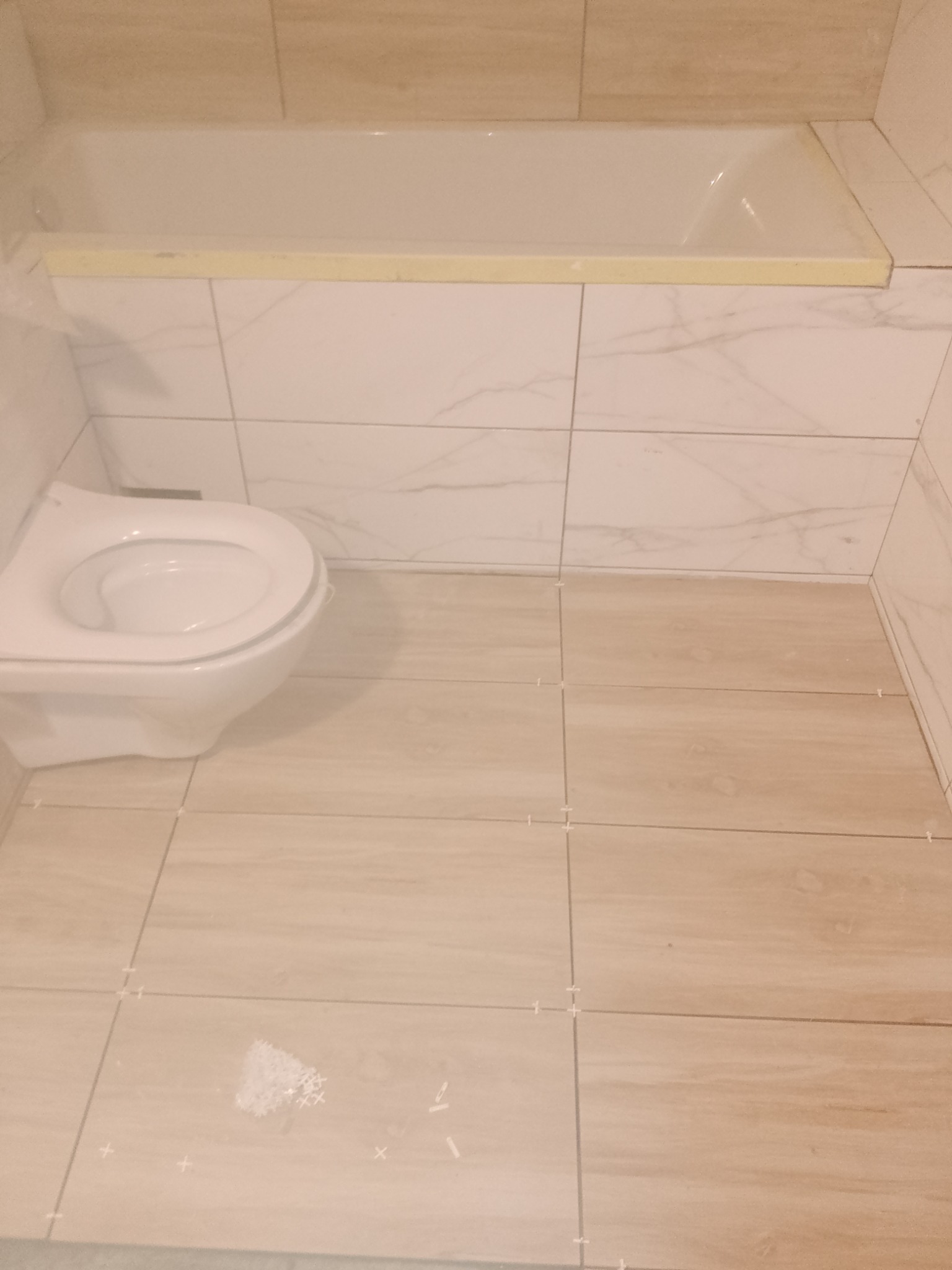 Układanie płytek w łazience, widoczna wanna z zabezpieczeniem, toaleta i podłoga w trakcie kładzenia płytek z krzyżykami dystansowymi, na podłodze pozostałości po kleju.