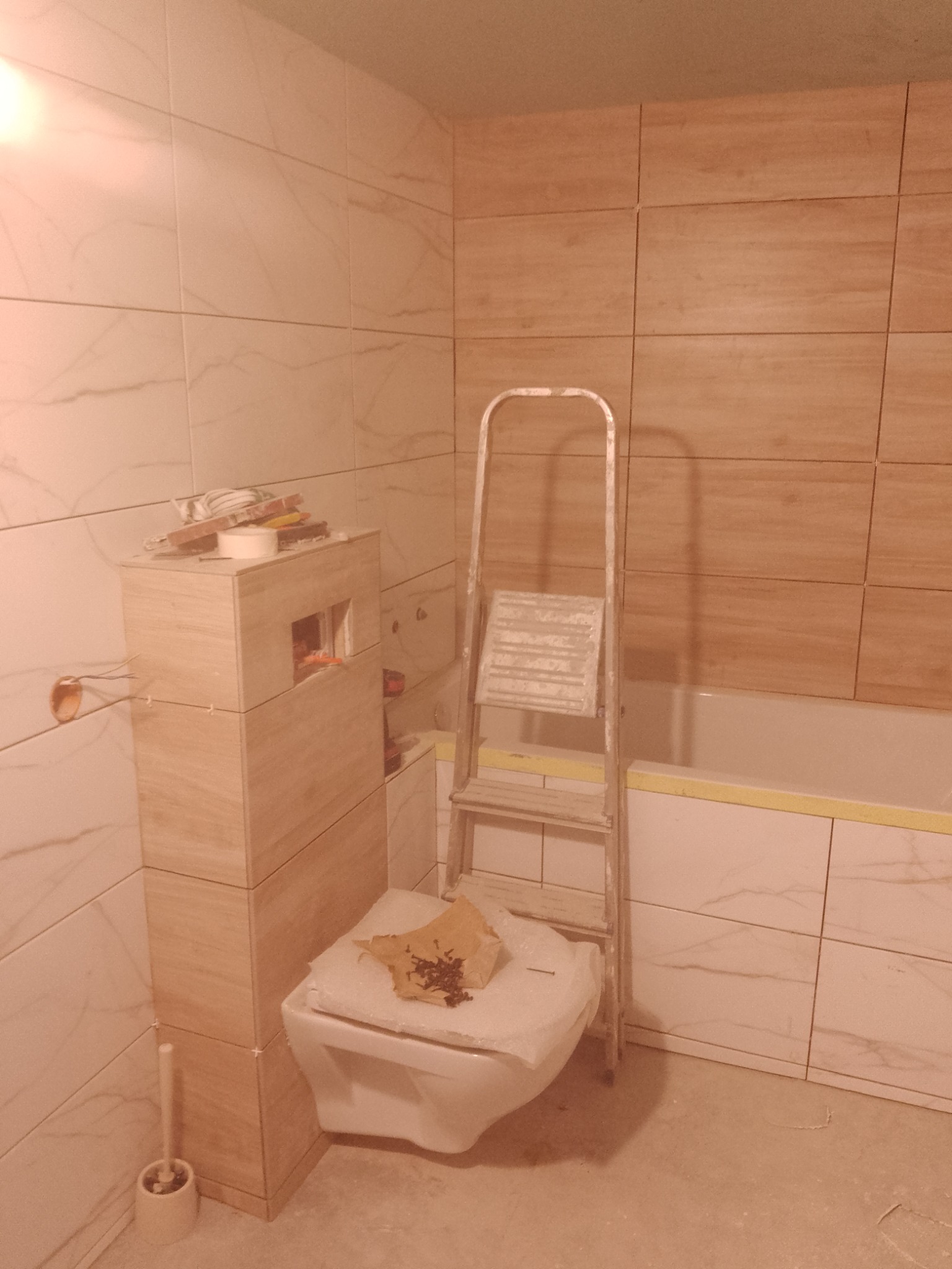 Łazienka w trakcie prac wykończeniowych: częściowo ułożone płytki imitujące drewno na ścianie, obudowa WC z płytek, drabina oparta o wannę, widoczne narzędzia i materiały budowlane.