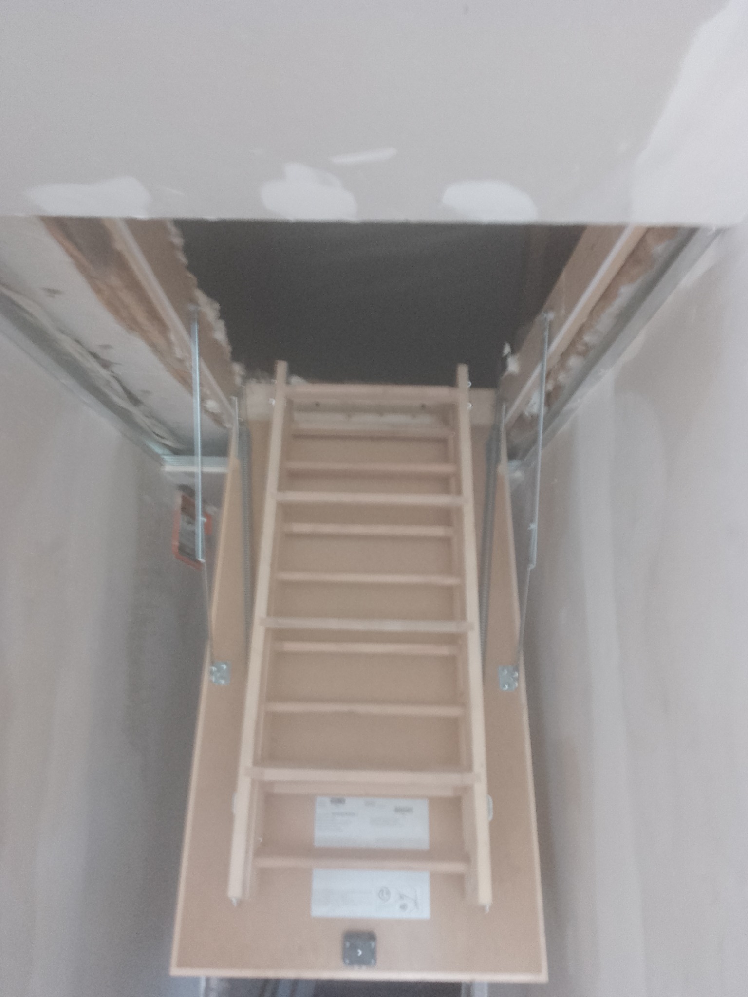 Zamknięte drewniane schody strychowe, widoczne mocowania do sufitu, ściany w trakcie szpachlowania, sufit z widocznymi łączeniami płyt gipsowo-kartonowych.