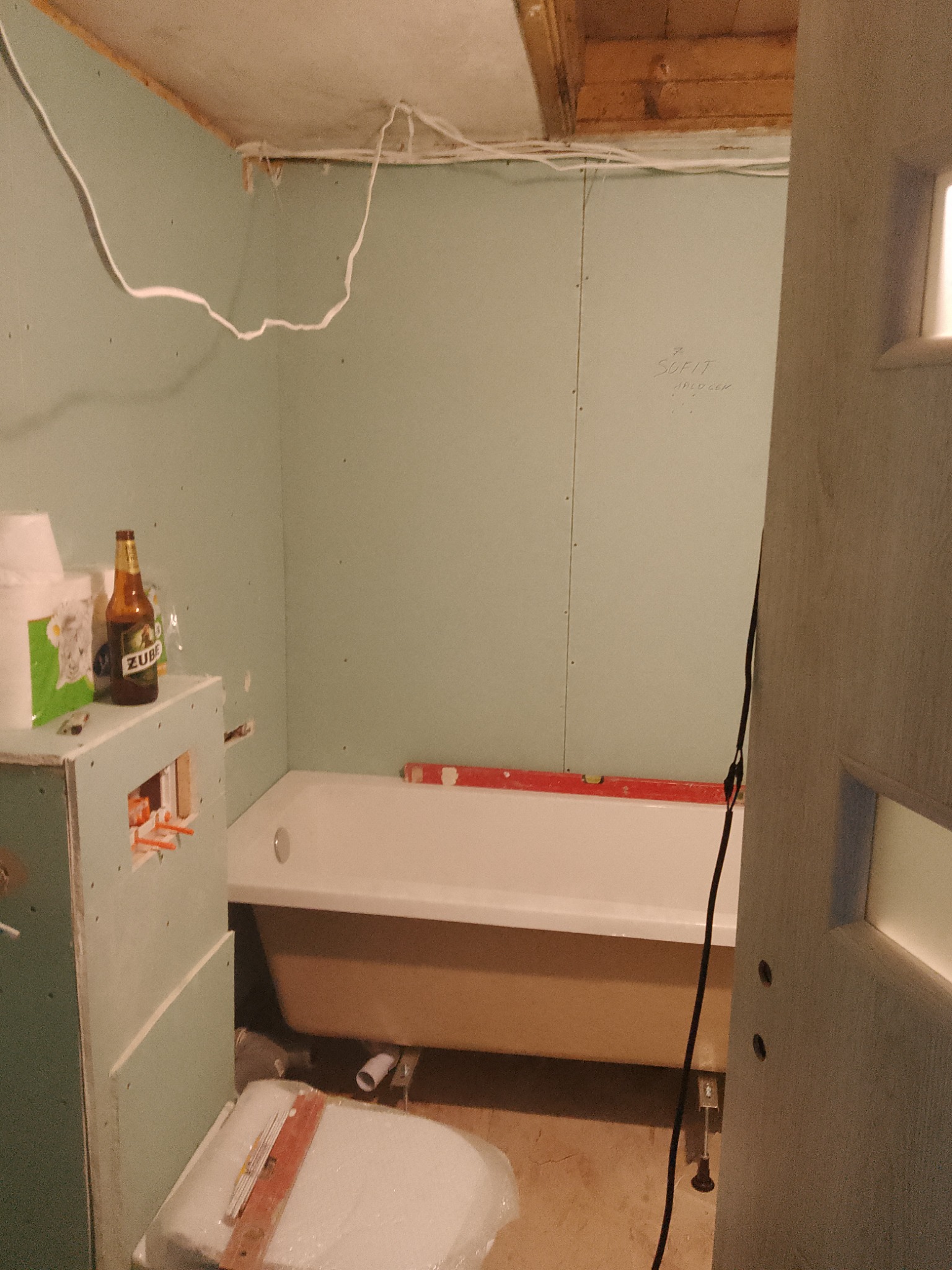 Łazienka w trakcie prac: wanna częściowo obudowana, ściany wyłożone płytami gipsowo-kartonowymi, widoczne przewody elektryczne, poziomica na wannie, butelka piwa na obudowie.