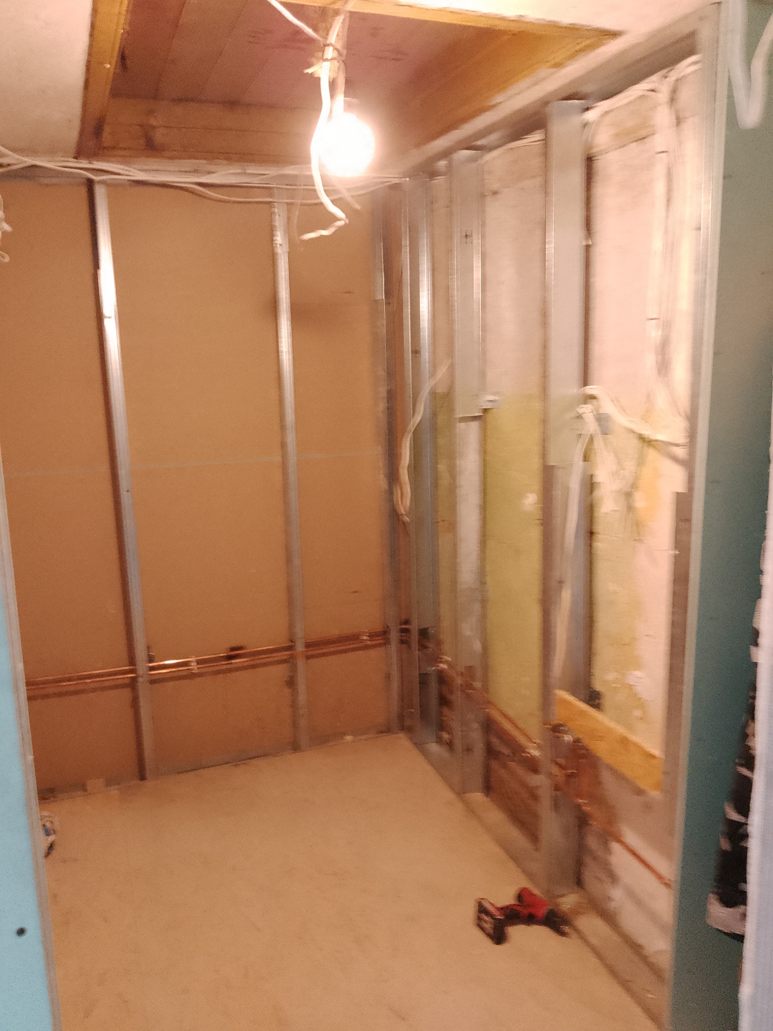 Wnętrze w trakcie prac przygotowawczych do wykończenia, widoczne metalowe profile, instalacja miedziana, fragment ocieplenia ściany, przewody elektryczne i wisząca żarówka.