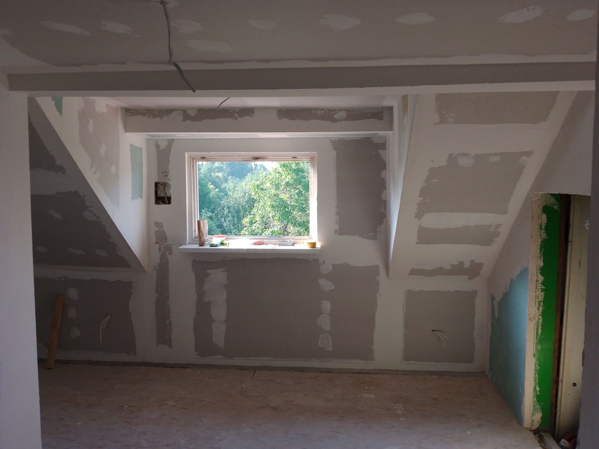 Poddasze w trakcie remontu, ściany pokryte płytami karton gips, widoczne łączenia do szpachlowania, okno z widokiem na zieleń.