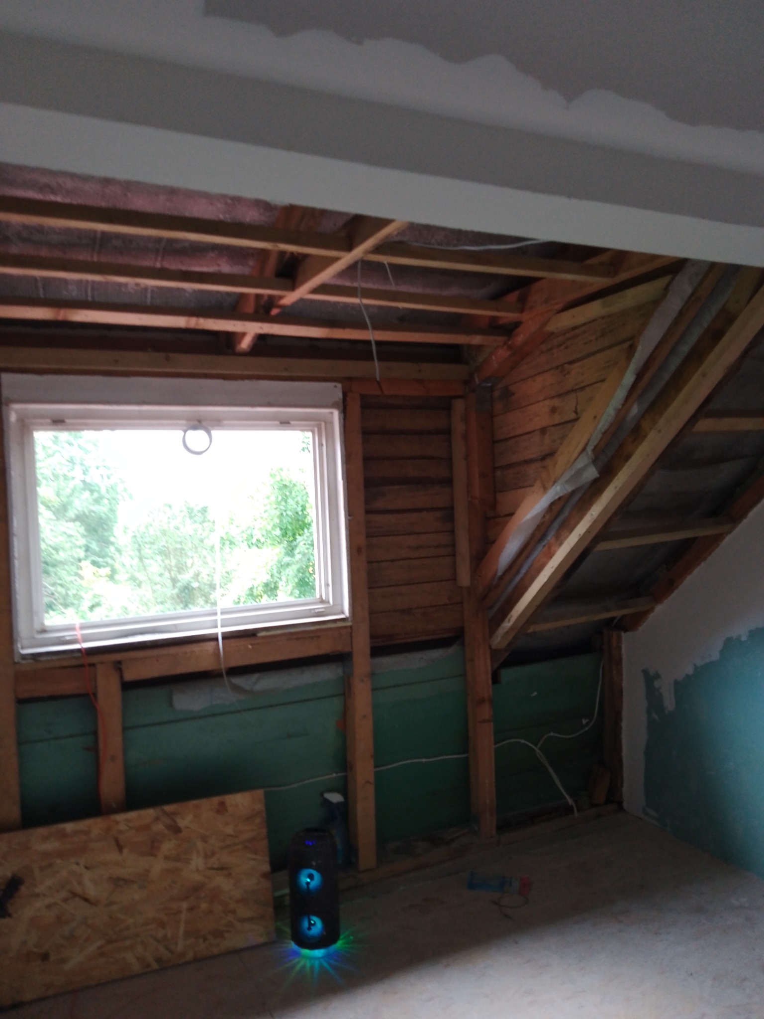Poddasze w trakcie adaptacji, widoczne drewniane krokwie, okno z widokiem na zieleń, częściowo otynkowana ściana i przenośny głośnik z podświetleniem LED na podłodze.