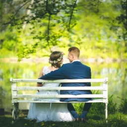 Para młoda siedzi na białej ławce z widokiem na jezioro, panna młoda ma wianek na głowie, a pan młody niebieski garnitur.