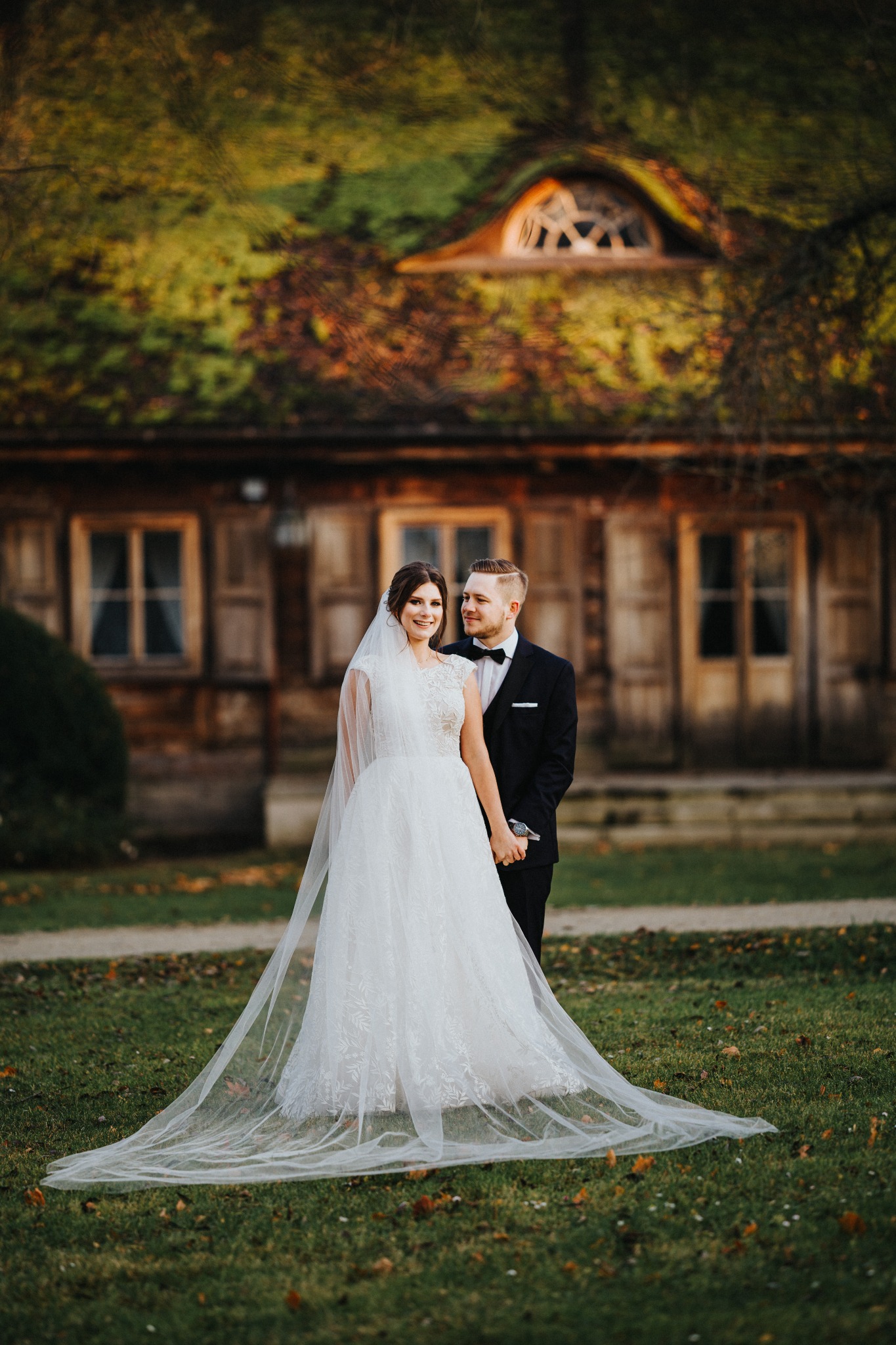 Para młoda, panna młoda w koronkowej sukni z długim welonem i pan młody w czarnym smokingu, pozują na tle drewnianego domu z oknami w stylu rustykalnym.