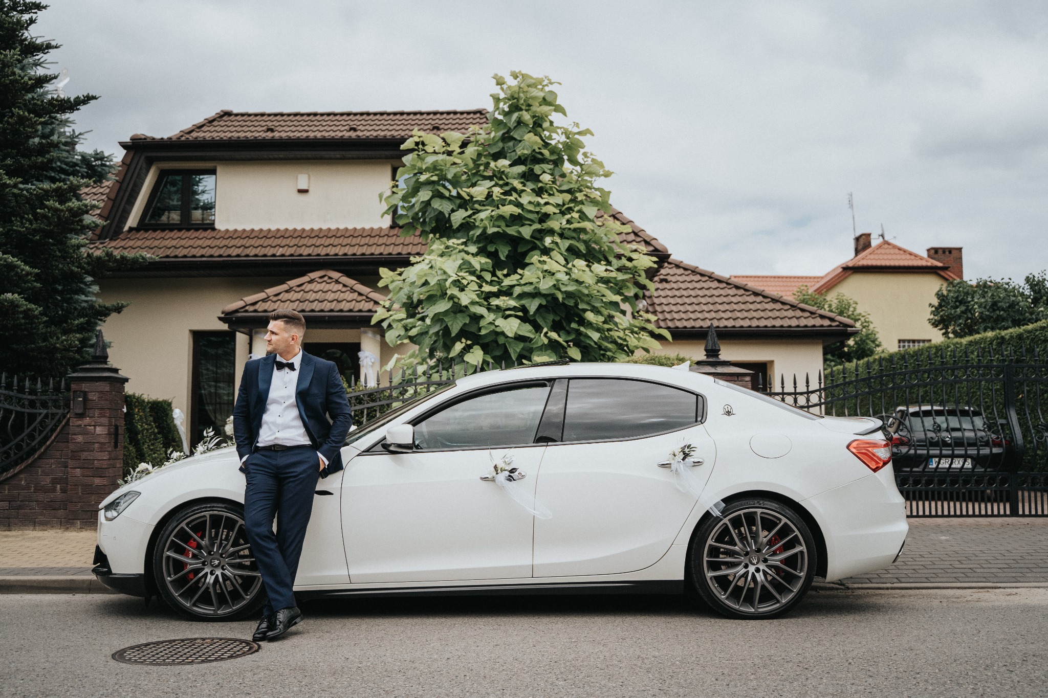 Elegancki mężczyzna w granatowym garniturze i muszce, oparty o białe, udekorowane kwiatami auto przed domem z brązowym dachem.